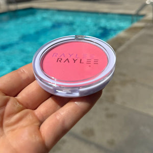 Hand holding a pink makeup compact with 'Raylee' branding by a poolside.